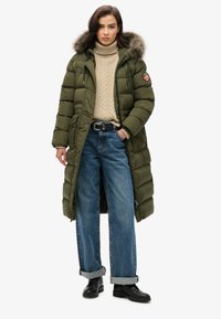 Long olive puffer coat with fur-lined hood, featuring a zip and button closure, worn over a beige knitted sweater and blue wide-leg jeans.