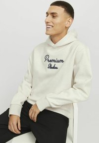 Light beige hoodie with a navy embroidered "Premium Studios" logo. Features a kangaroo pocket and ribbed cuffs, styled with black pants.