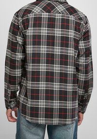 Camisa de franela a cuadros en blanco y negro con acentos rojos, de mangas largas y textura tejida. Presenta un patrón a cuadros y puños con botones.