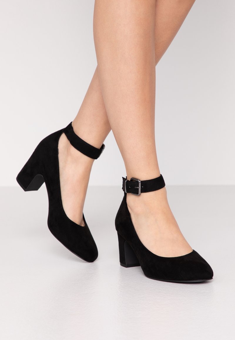 Femme portant des talons carrés en daim noir avec bride à la cheville et bout arrondi, debout sur un sol clair devant un fond uni.