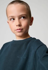 Tamsiai žalias striukinis megztinis su tekstūra, raukšlėtu audiniu, apvalia iškirpte ir siuvinėta "ALLSAINTS" logotipu krūtinėje. Lygus audinio paviršius.