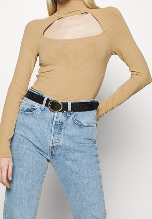 Long-sleeve, high-neck top in light brown ribbed fabric with square cutouts at shoulders, paired with high-waisted light blue jeans and a black belt.