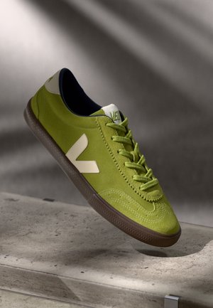 Green suede sneaker with a white logo and accents. Features round toe, textured brown rubber sole, and flat laces.