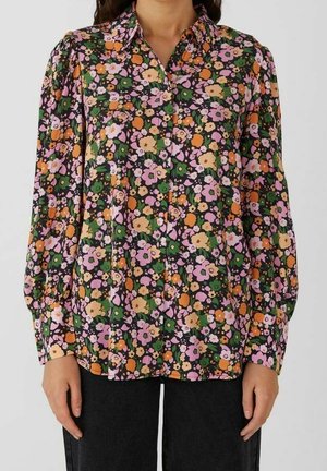 Kvinde iført en langærmet skjorte med knapper og et blomstermønster i orange, pink, gul og grøn, kombineret med sorte bukser.