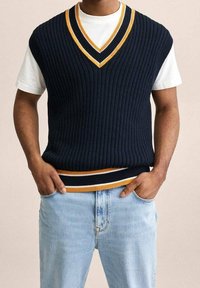 Marineblauer, gerippter Strickpullover ohne Ärmel mit V-Ausschnitt, versehen mit gelben Akzenten am Kragen und Saum, kombiniert mit einem weißen T-Shirt und hellen Jeans.
