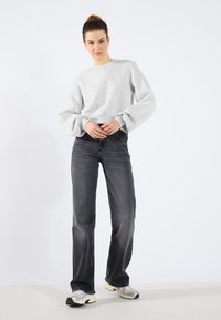 Felpa grigia con maniche a costina, abbinata a jeans neri. Il modello indossa sneakers grigie e nere. Lo sfondo chiaro mette in risalto l'outfit.