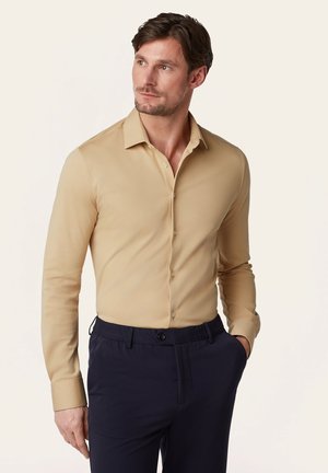 Man in een beige overhemd met lange mouwen en donkere marineblauwe broek, staand met één hand in de zak, kijkt opzij.