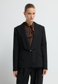 Zwarte op maat gemaakte blazer met een geknipt revers, sluiting met één knoop en een streepjespatroon. Gecombineerd met een bruine blouse met een strikdetail.