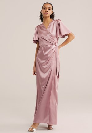 Ženska s kodrastimi lasmi, ki nosi biserne uhane in vijolično satenasto ovijalno obleko z razširjenimi rokavi ter srebrnimi odprtimi čevlji s peto, stoji z roko na boku.