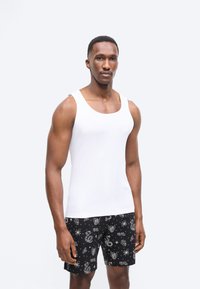 Άνδρας που φοράει λευκό αμάνικο τοπ και μαύρο σορτς με μοτίβα λευκού φιδιού, ιστού αράχνης και έμβλημα, στέκεται μπροστά σε απλό φόντο.