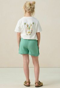 Witte korte mouwen t-shirt met kreeftafbeelding op de rug, gecombineerd met groene gestructureerde shorts. Bruine sandalen maken de outfit af.