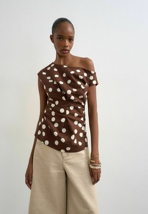 Femme portant un top marron asymétrique à pois blancs et un pantalon beige à jambes larges, accessoirisée avec plusieurs bracelets.
