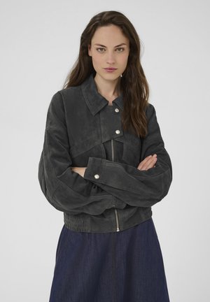 Woman with long brown hair wearing a dark gray suede jacket with zipper and silver buttons, arms crossed, and a dark blue skirt.