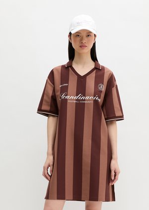 Vrouw die een bruin gestreepte korte mouw voetbalstijl jurk draagt met de tekst "Scandinavia Football Community" en een witte pet.