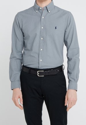 Mężczyzna noszący szarą koszulę na guziki z małym granatowym logo na piersi, czarne spodnie i czarny pasek z srebrną klamrą.