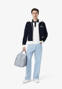 Chaqueta azul marino con cremallera y cuello crema, polo blanco con boutonera azul marino, jeans claros y bolso estructurado gris. Zapatillas blancas completan el look.