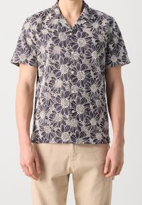 Homme portant une chemise à manches courtes boutonnée avec un grand motif floral de marguerites noir et blanc, portée sortie sur un pantalon beige.