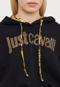Felpa nera con testo "just cavalli" in oro borchiato, caratterizzata da un cappuccio interno giallo a motivo e lacci. Il tessuto appare liscio e spesso.