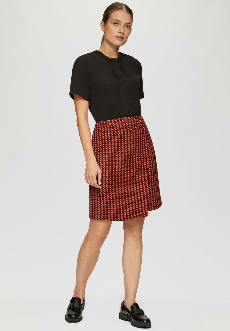 Zwart T-shirt met korte mouwen en een geplooide halslijn; rok met een rood patroon en een houndstooth ontwerp; gedragen met zwarte instappers.