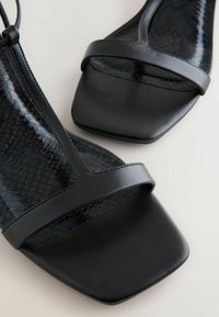 Sandalias negras con un diseño de punta cuadrada, una correa de cuero suave y un panel negro con textura de reptil.