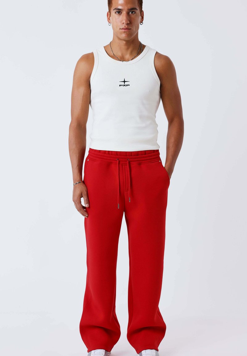 Jeune homme portant un débardeur blanc avec le logo "PXP" et un pantalon de survêtement rouge ample avec cordon de serrage, debout face à nous, les mains dans les poches.