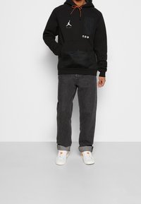 Mężczyzna ubrany w czarną bluzę Nike Jordan z kieszeniami z przodu, ciemne dżinsy z wywiniętymi nogawkami i białe trampki z pomarańczowymi akcentami, stoi przy szarej ścianie.