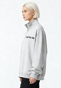 Grijze half-zip trui met een comfortabele pasvorm, voorzien van lange mouwen en zwarte tekst "C'est la vie" op de borst.