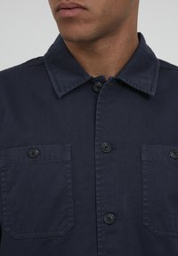 Camicia blu navy con colletto, dotata di due taschini sul petto, bottoni neri e una trama di tessuto robusta.