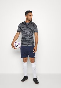 Grijze camouflage sportshirt met korte mouwen, gecombineerd met marineblauwe shorts. Het model houdt een witte bal vast met roze en paars accent. Zwarte sportschoenen en witte sokken.