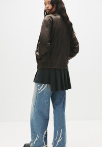Giacca bomber in pelle marrone con vita elastica, indossata sopra una gonna plissettata nera. Abbinata a jeans azzurri chiari con macchie di vernice bianca.