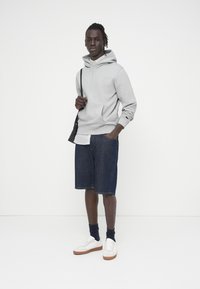 Sweat à capuche gris clair avec une poche kangourou, associé à un short en jean foncé et des baskets blanches avec des semelles marron. Sac à bandoulière noir visible.