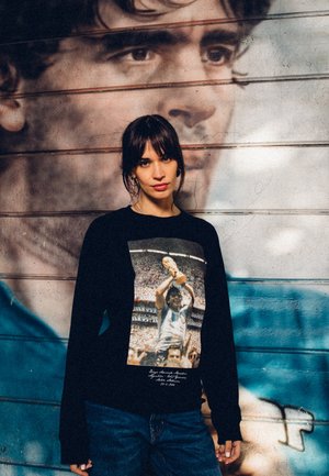 Zwart langemouwen shirt met een kleurafdruk van een voetballer met een trofee, tegen een gedempt muurschildering. De stof lijkt glad te zijn.