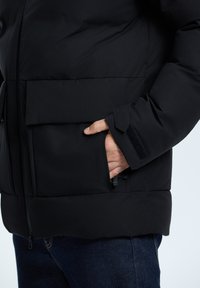 Musta puuvillapuku, jossa on vetoketjullinen etuosa, suuri rintatasku ja tarranauha korostaa hihoja. Liukas materiaali, jossa on pehmeä koostumus.