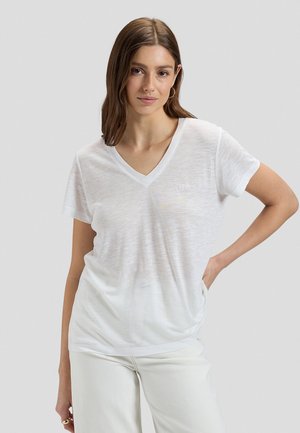 Frau mit schulterlangen braunen Haaren, die ein weißes V-Ausschnitt-T-Shirt und weiße Hosen trägt, posiert mit einer Hand in der Hüfte.