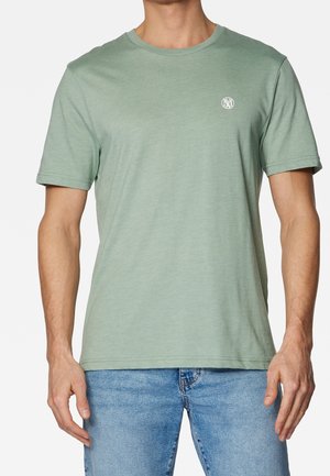 Mann in einem einfarbigen hellgrünen T-Shirt mit kurzen Ärmeln und einem kleinen weißen Logo auf der linken Brust, kombiniert mit hellblauen Jeans.