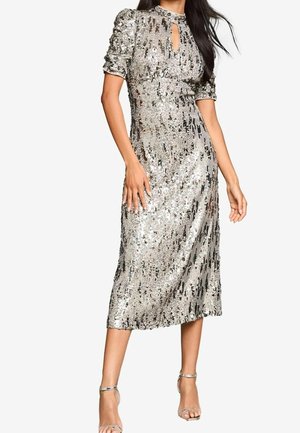 Mujer que lleva un vestido midi plateado con lentejuelas, mangas cortas abullonadas y escote con abertura, combinado con zapatos de tacón plateados con correa en el tobillo.
