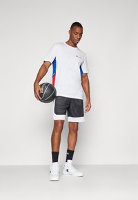 Conjunto atlético masculino: camiseta blanca con paneles laterales azules y rojos, pantalones cortos de malla negra con ribetes blancos, sosteniendo un balón de baloncesto, zapatillas blancas.