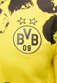 Κίτρινη αθλητική φανέλα με μαύρο λογότυπο "BVB 09" και δύο αστέρια από πάνω, με αφηρημένο μαύρο μοτίβο στο πάνω μέρος.