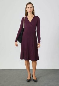 Γυναίκα που φοράει ένα μωβ φόρεμα με μακριά μανίκια, πλισέ, έως το γόνατο, με μαύρες γόβες, χρυσούς κρίκους και μια μαύρη τσάντα ώμου.