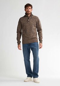 Brun, texturerad stickad tröja med hög, ribbad krage och treknappslå, kombinerad med blå jeans och vita sneakers.