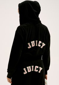 Zwarte velours sportoutfit met een capuchon en "JUICY" in roze en groene borduurrand aan de achterkant. Zachte textuur en losse pasvorm met een aantrekkoord in de taille.