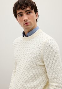 Pullover aus cremefarbenem, strukturierter Strick mit Rundhalsausschnitt und Diamantmuster, getragen über einem hellblauen Hemd mit Kragen. Weicher Stoff, lässiges Design.