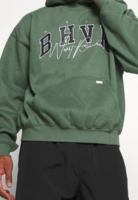 Zielona bluza z kapturem, z przednią kieszenią i wyrazistym nadrukiem z napisem "BHV" oraz "Worst Behavior" w kontrastujących kolorach.