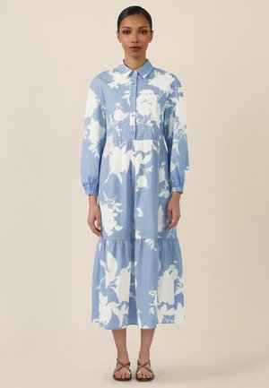 Frau trägt ein blaues Midikleid mit langen Ärmeln und weißem Blumenmuster, Knopfleiste vorne, geraffter Taille und beige flache Sandalen.