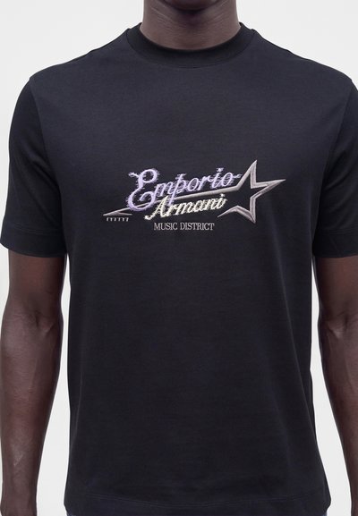 Μαύρο T-shirt με το κείμενο "Emporio Armani Music District" και σχέδιο αστέρι κεντημένο στο στήθος, που φοριέται από άτομο με σκούρο δέρμα.