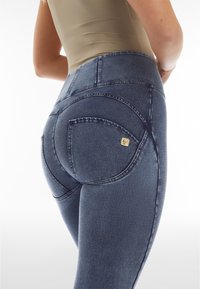 Leggings de mezclilla de alta cintura en azul oscuro. Presentan un diseño ajustado, costuras visibles, bolsillos traseros curvados y un pequeño logo de acento en dorado.