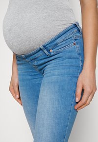 Jeans de maternidad en denim azul claro, con un diseño ajustado, una cintura ligeramente más alta y una textura suave. Los detalles incluyen remaches de cobre.