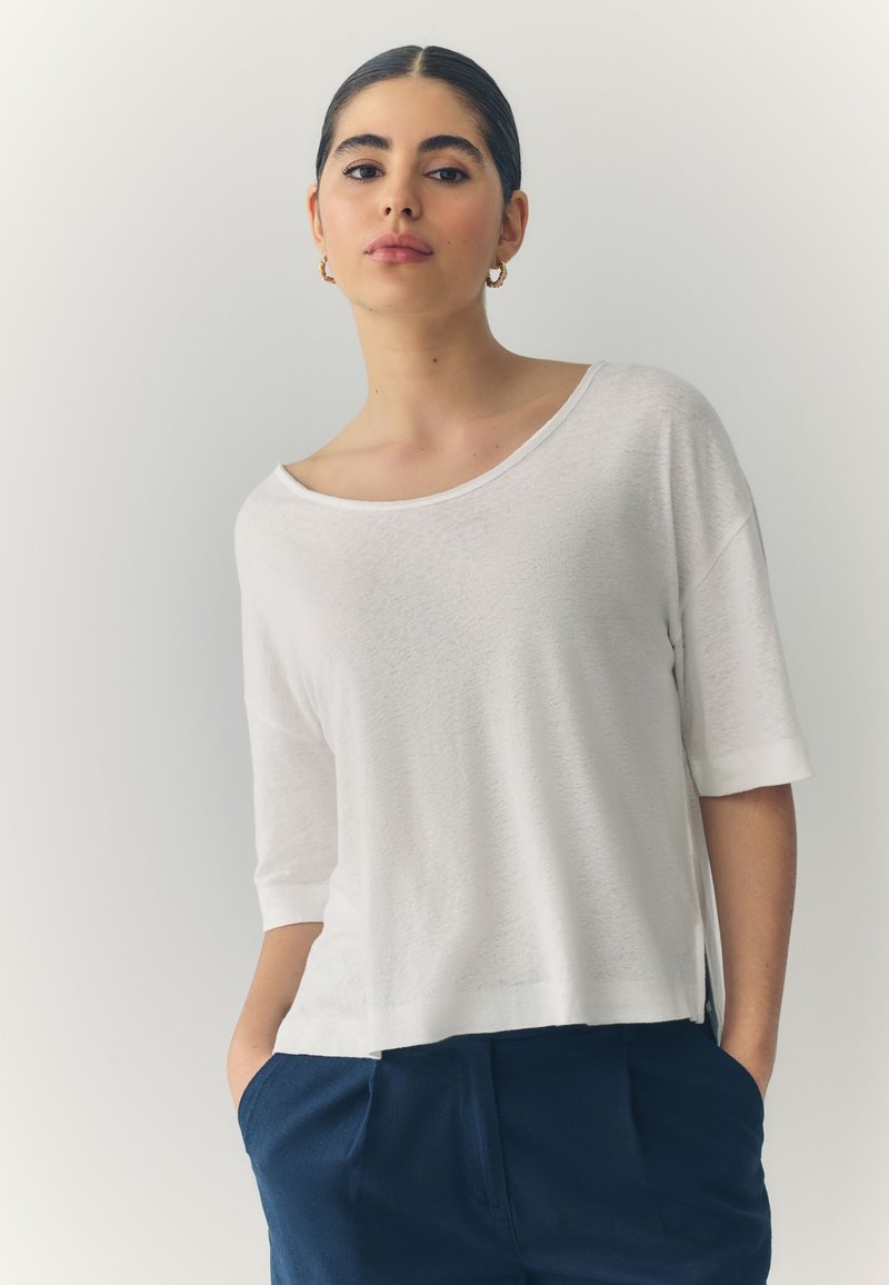 White, loose-fitting short-sleeve top with a wide neckline and soft texture, paired with dark blue trousers. Notable side slits for ease.
