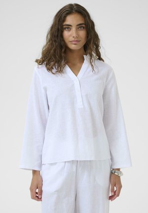 Kobieta o kręconych włosach, ubrana w luźną białą koszulę z długim rękawem i guzikami oraz dopasowane białe spodnie, stojąca na jednolitym tle.