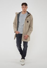 Giacca impermeabile beige con interni grigi, tasca frontale e cappuccio regolabile, abbinata a una maglietta grigia e jeans scuri. Sneakers bianche completano l'outfit.